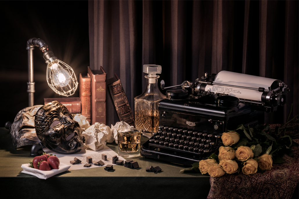 ﻿Still life photograph of typewriter, masks, flowers, and books on a desk